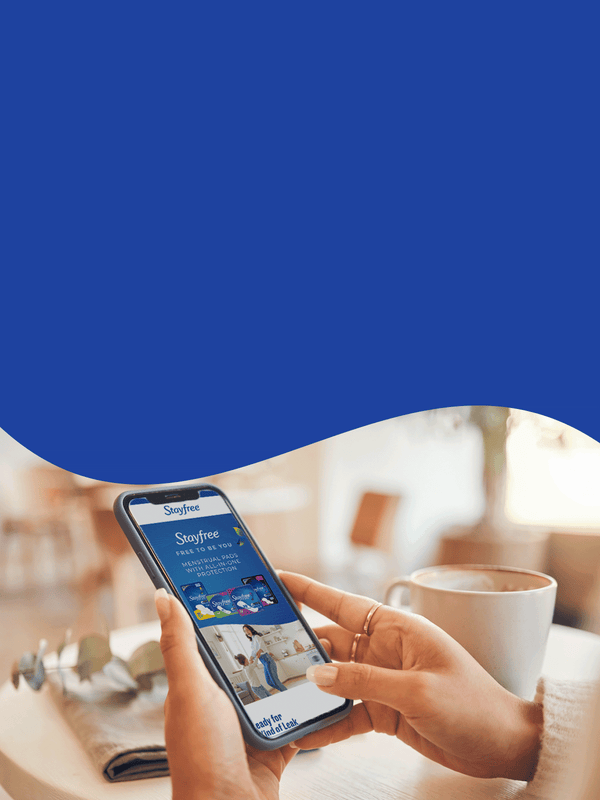 Person holding a smartphone with Stayfree website open, sitting at a table with a coffee cup.